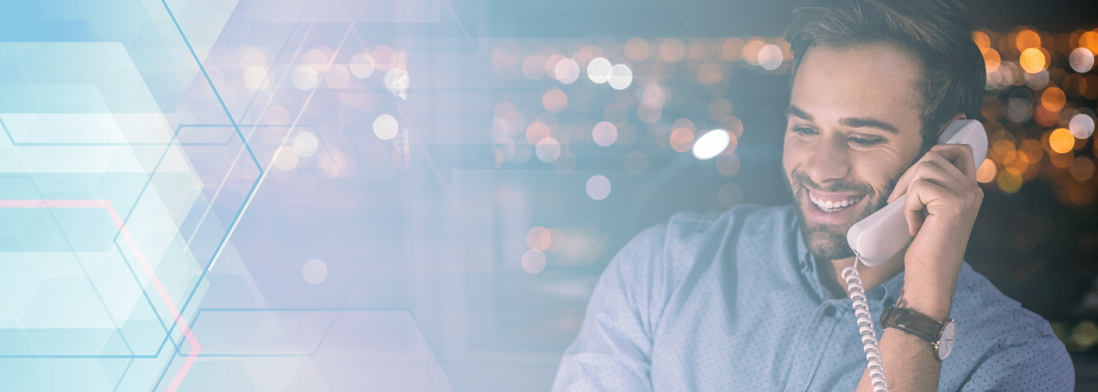 a young male professional smiles while talking on a SIP phone. He has brown hair, light skin and wears a blue collared shirt